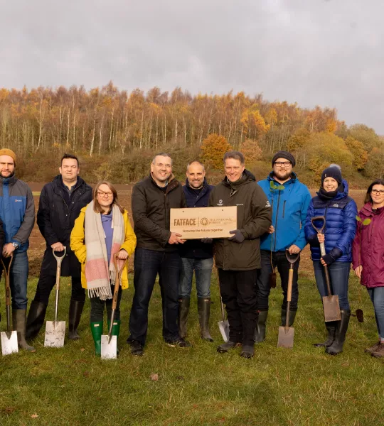 A group of people, many holding spades, are standing in a field with a mature woodland in the background. The three in the middle are holding a wooden plaque with the FatFace and National Forest Company logos on.